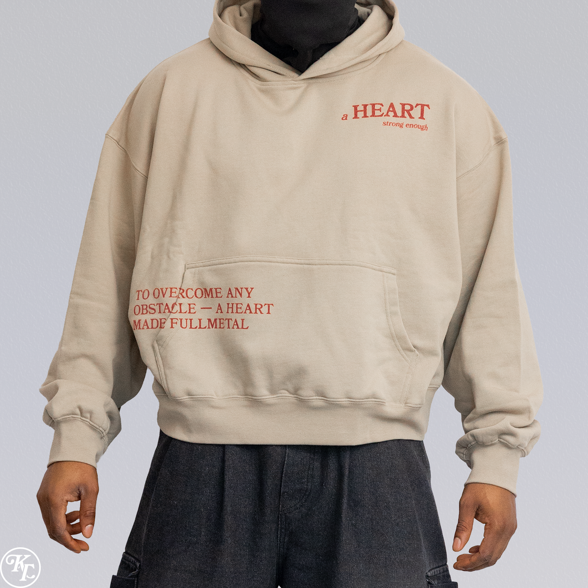 A person wearing a beige pullover hoodie with red printed text on the chest and front pocket area, paired with dark pants, against a light gray studio background.
