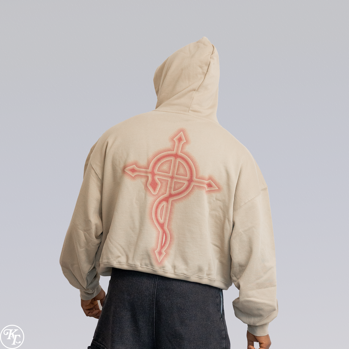 A person shown from the back wearing a cropped beige hoodie with a large pink glowing cross graphic on the back, paired with dark gray pants, against a light gray studio background.