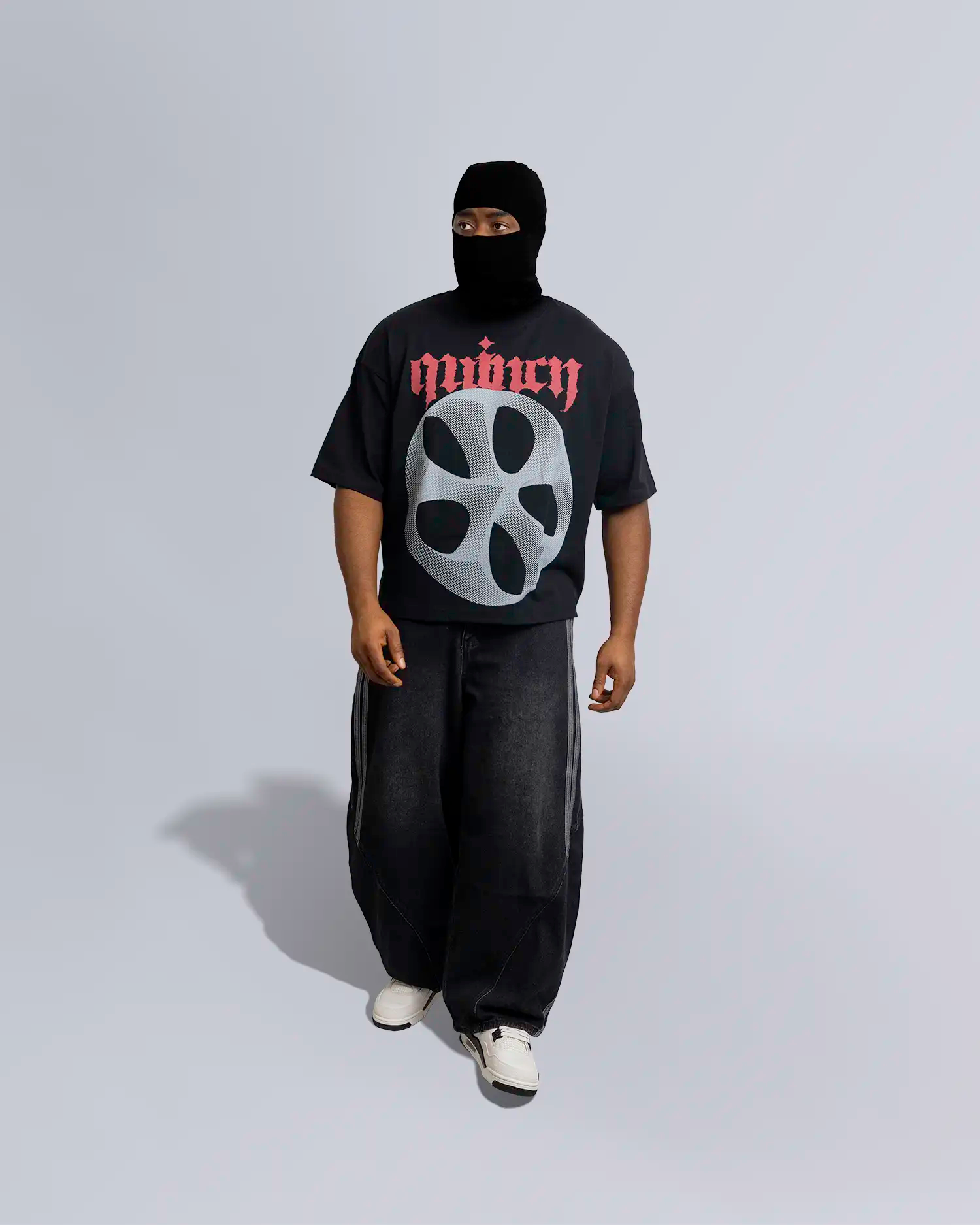 Person wearing a black t-shirt with a logo and text, dark pants, and white shoes on a light gray background